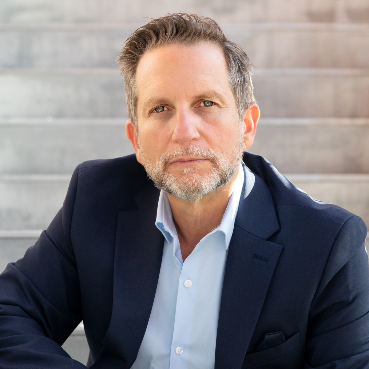 A bearded man in a suit and light blue shirt sits on steps, looking directly at the camera with a serious expression.