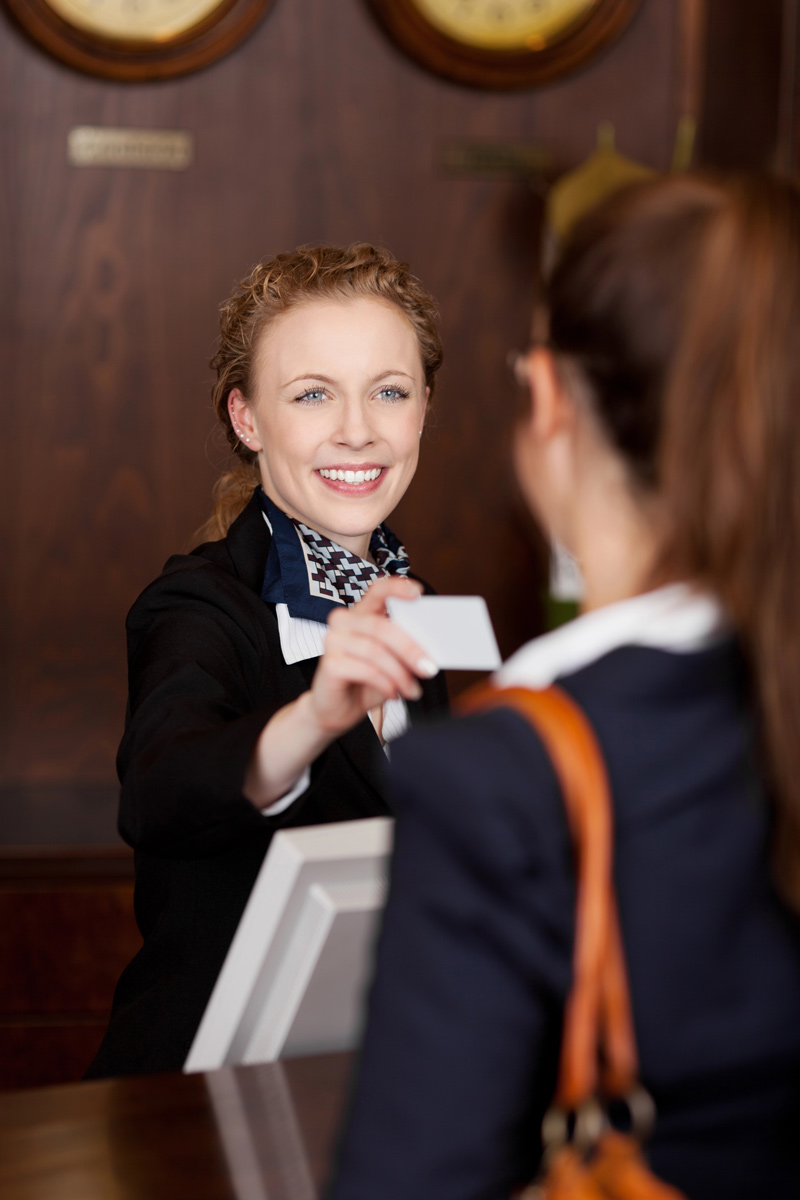 A smiling hotel receptionist hands a card to a guest at the front desk, set against a backdrop of wooden decor and wall clocks.