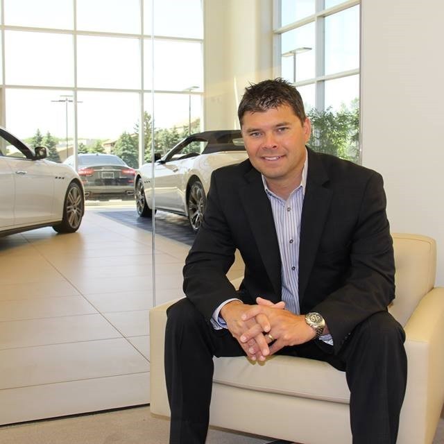 A man in a suit sits on a chair in a car dealership, with luxury cars visible in the background and ample natural light streaming in.