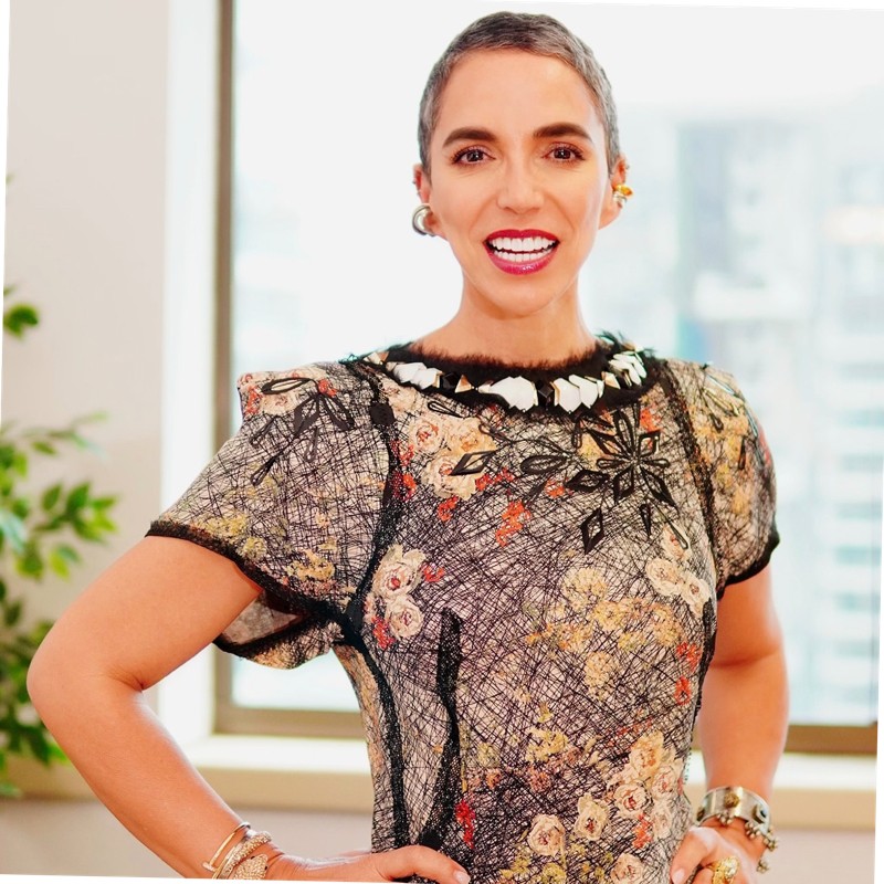 A woman in a floral dress poses confidently, smiling brightly in a bright indoor setting, with a cityscape visible through the window behind her.
