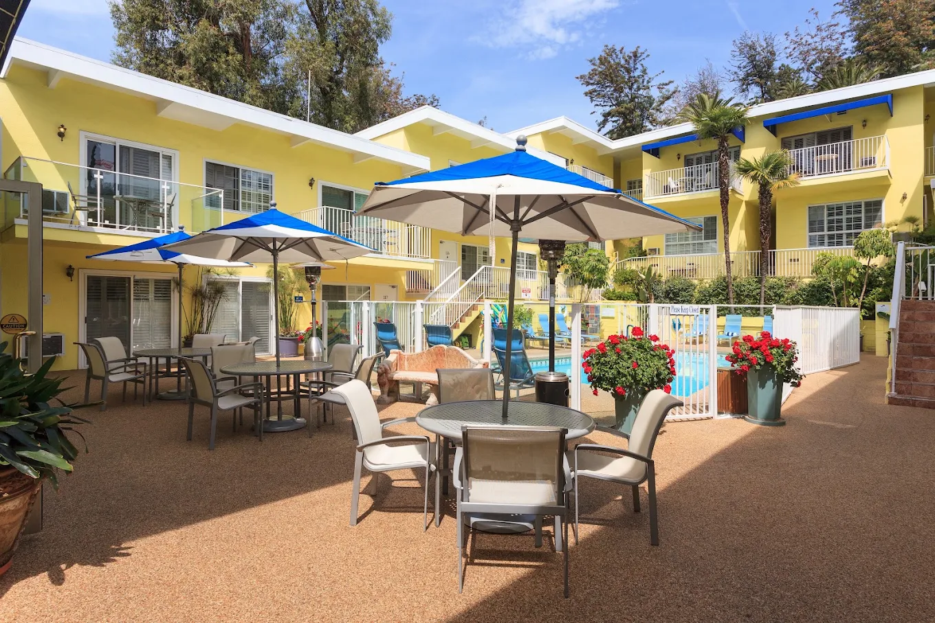 The image shows a sunny courtyard with yellow buildings, blue umbrellas over tables, and a swimming pool surrounded by greenery and flowers.